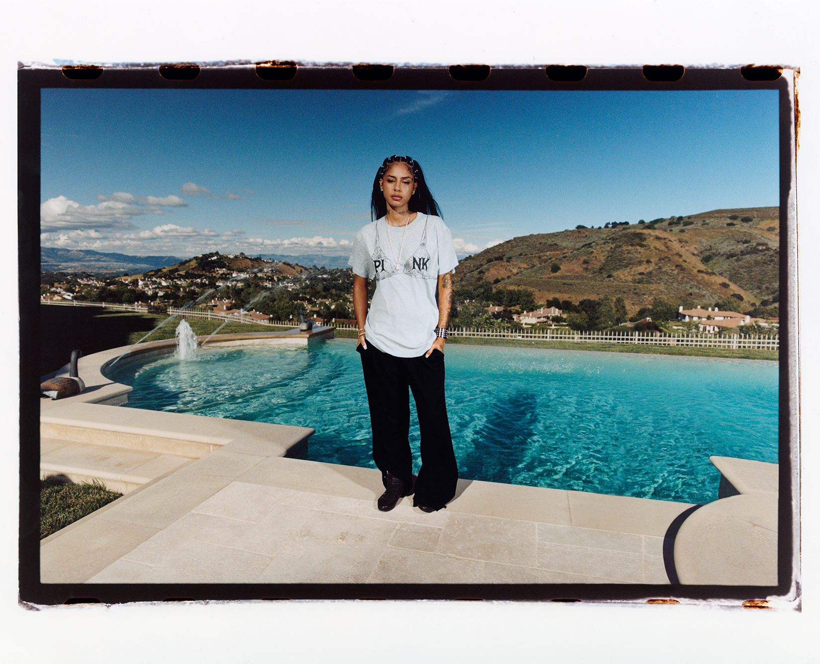 Model in a while t shirt and black pants posing with her hands in her pocket in front of a pool on a sunny day